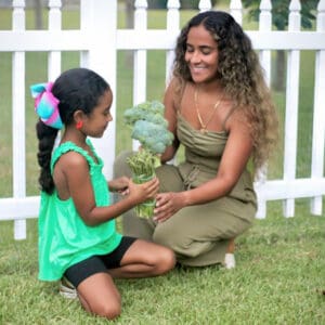 Woman and girl holding broccoli by white fence.