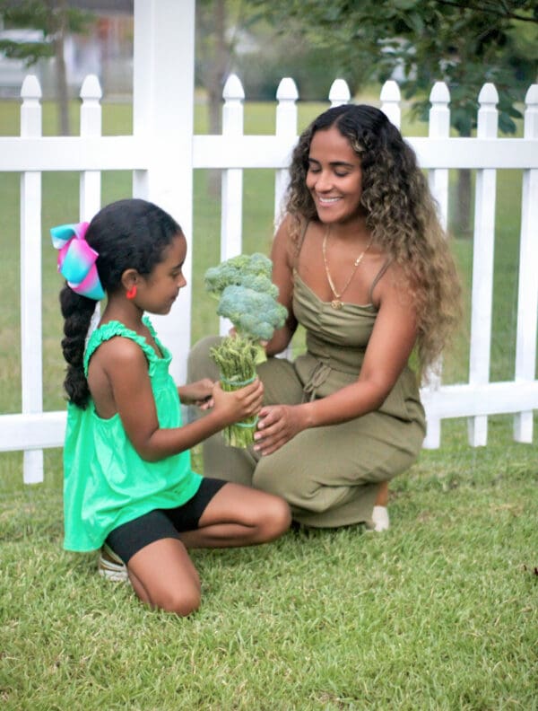 Woman and girl holding broccoli by white fence.